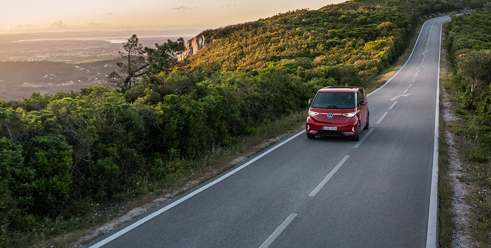Roter Volkswagen ID. Buzz f&auml;hrt auf einer kurvigen Landstra&szlig;e durch eine gr&uuml;ne H&uuml;gellandschaft bei Sonnenuntergang &ndash; Symbol f&uuml;r Freiheit, Nachhaltigkeit und moderne Mobilit&auml;t.